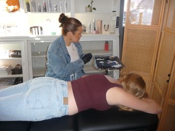 Cosmetic technician in denim jacket and gloves prepares instruments on a tray while a client lies face down on a treatment table in a cozy beauty salon room with product shelves and a wicker privacy screen.