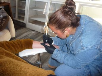 Focused tattoo artist in a denim jacket and black gloves tattooing a client's forearm on a padded armrest in a bright tattoo studio.