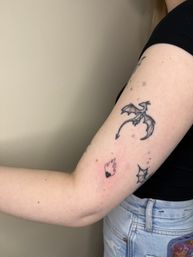 Close-up of upper arm tattoos on light skin: small black-ink flying dragon, flaming heart, stars and spiderweb, worn with a black tank top and light denim jeans against a neutral wall.