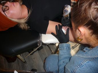 Close-up in a tattoo studio of an artist in black gloves using a plastic-wrapped tattoo machine to ink a client's inner forearm while the client reclines on a black chair, wearing glasses and an orange beanie.