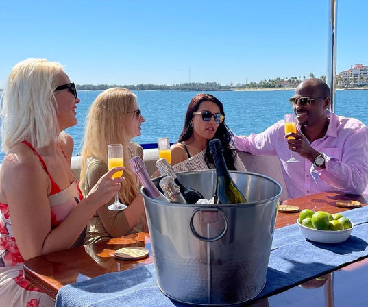 Four adults enjoying mimosas on a sunny yacht, champagne bucket and bowl of limes on the table with blue ocean and palm-lined coastline in the background.