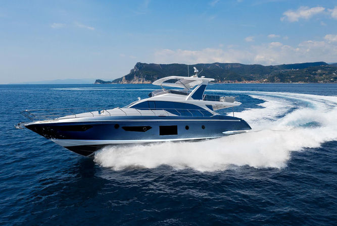 Sleek blue-and-white luxury motor yacht speeding across deep blue sea, carving a white wake with rocky coastline and clear sky in the background.