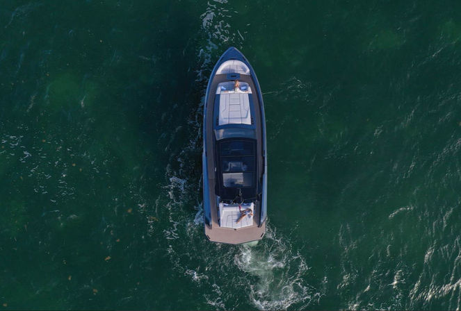 Aerial drone view of a sleek motorboat cruising emerald-green coastal waters, two people relaxing on sun pads at bow and stern with the boat’s wake trailing behind.