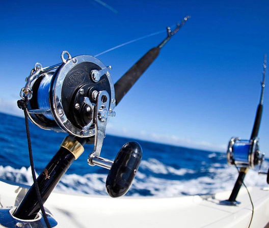 Offshore deep-sea fishing rods with chrome reels mounted on a boat, line trailing into the vibrant blue ocean under a clear sky