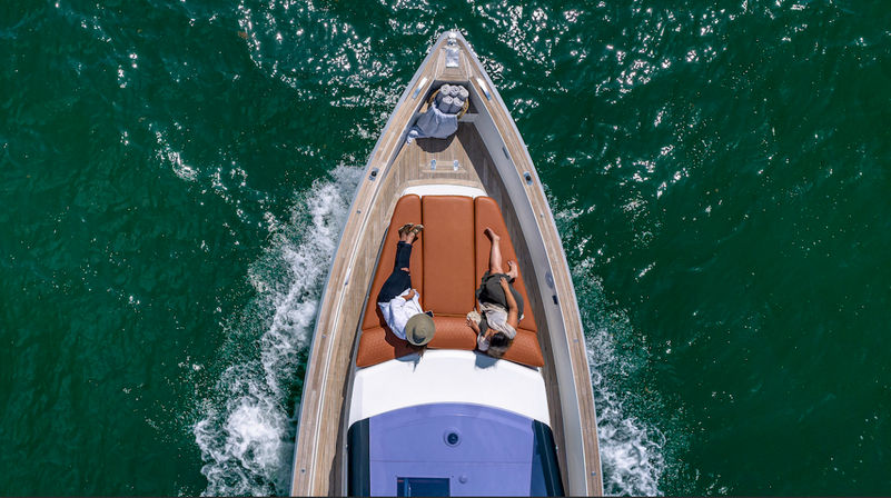 Aerial drone view of two people relaxing on the bow of a motorboat cruising emerald-green water on a sunny day