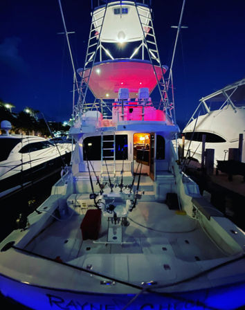 Nighttime sportfishing yacht docked at a marina, aft deck lined with fishing rods, tall tuna tower and blue-pink LED lighting.