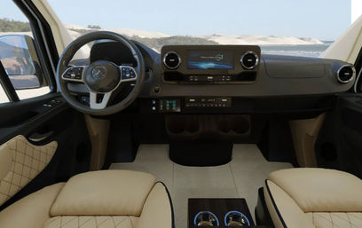 Stylish beige leather camper-van cockpit with modern dashboard and touchscreen, steering wheel, quilted door panels, center console cupholders, beach and sand dunes visible through the windshield.