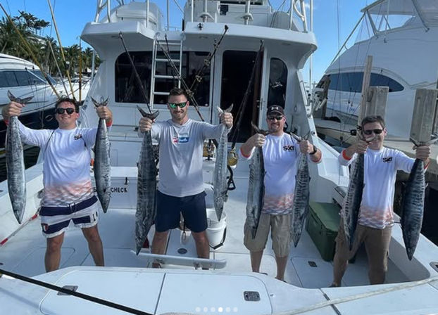 Four anglers on a sportfishing boat at a marina proudly holding long wahoo fish, with fishing rods, nearby yachts, and palm trees under a sunny sky.