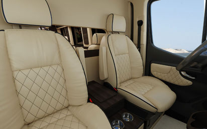 Luxury camper van interior with cream quilted leather captain seats and black piping, wooden center console with cup holders, and a sandy coastal view visible through the passenger window.
