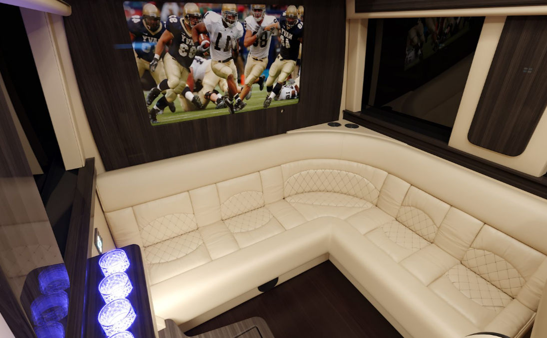 Cozy luxury RV lounge with cream quilted L-shaped leather sofa, dark wood paneling, wall-mounted TV showing a football game, LED-lit cup holders and ambient lighting.