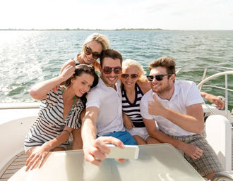 Five friends taking a cheerful selfie on a sunny boat cruise in coastal waters, wearing sunglasses with the sea and horizon in the background.