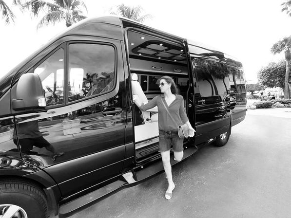 Woman in sunglasses stepping out of a black luxury shuttle van on a sunny driveway, palm trees reflected on the vehicle suggesting a tropical resort transfer.
