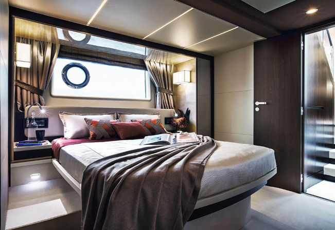 Modern luxury yacht cabin with round porthole ocean-view window, plush queen bed with patterned pillows and draped gray throw, bedside lamp and steps to the entrance.