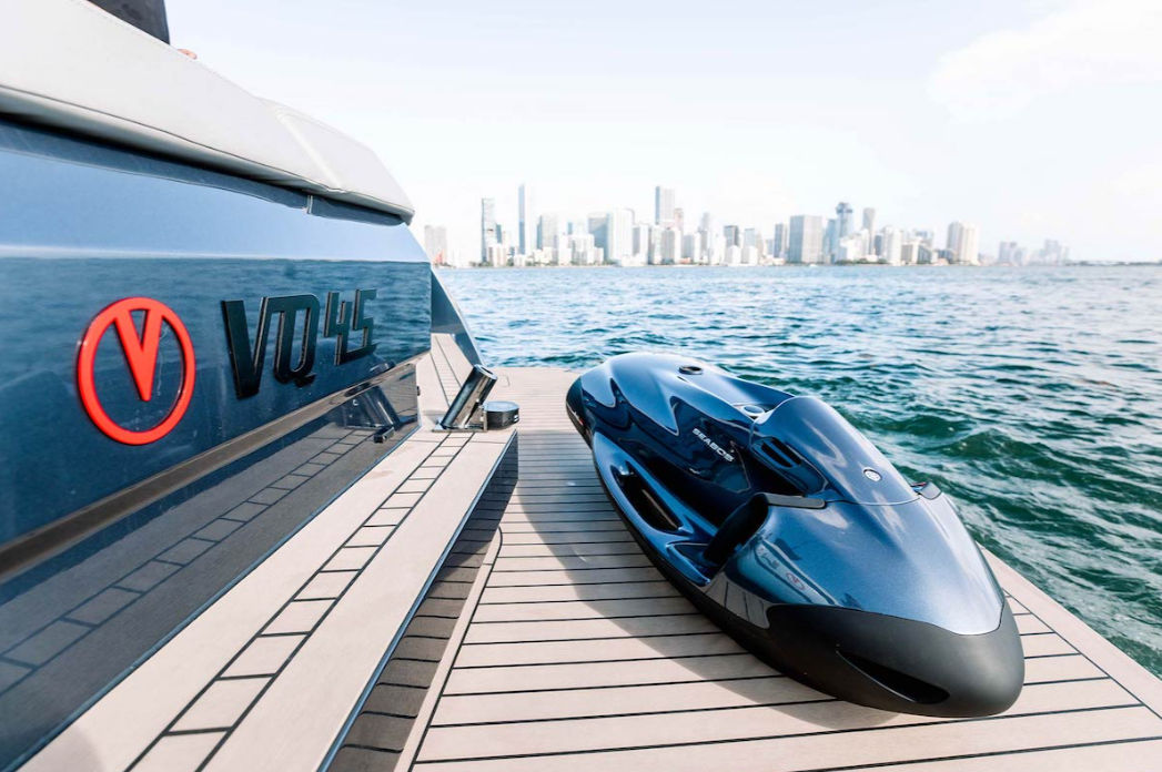 Sleek metallic blue personal watercraft resting on a yacht's swim platform with calm ocean water and a distant city skyline on a sunny day.