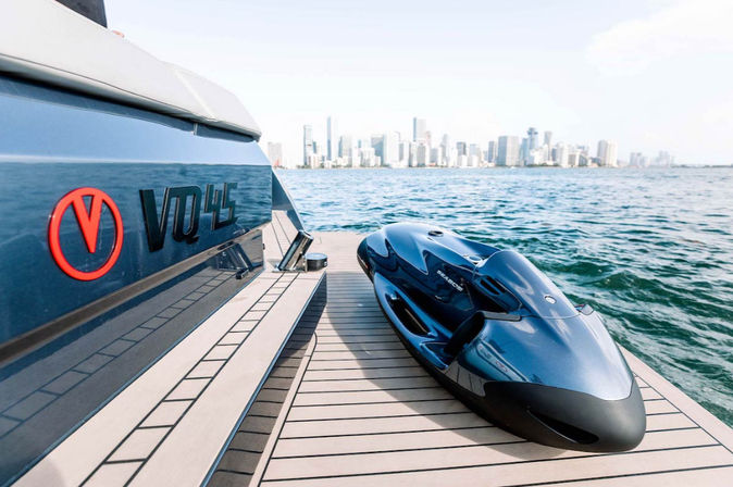Sleek metallic blue personal watercraft resting on a yacht's swim platform with calm ocean water and a distant city skyline on a sunny day.
