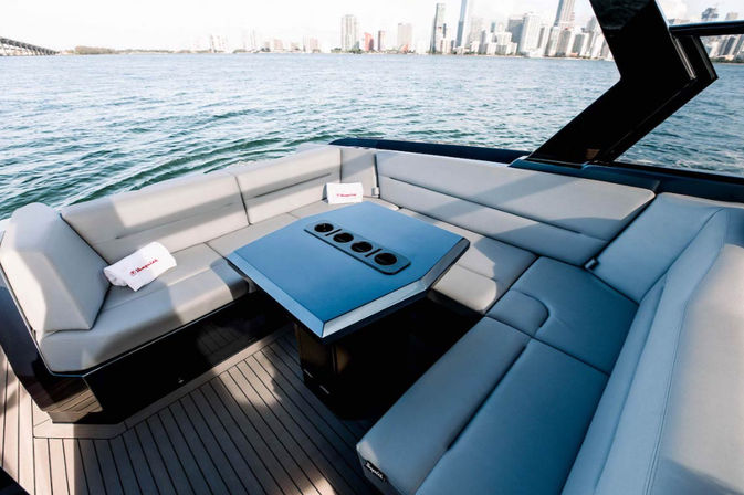 U-shaped gray leather lounge on a luxury yacht aft deck with a central table and cup holders, overlooking blue harbor water and a distant urban skyline