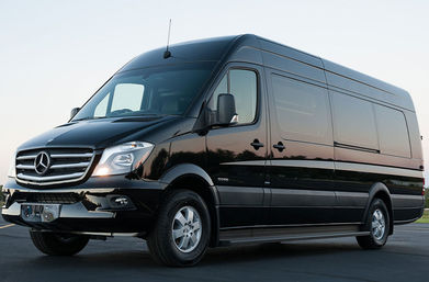Sleek black Mercedes passenger van parked on asphalt at dusk, front-side view showing grille, headlights and long cargo cabin