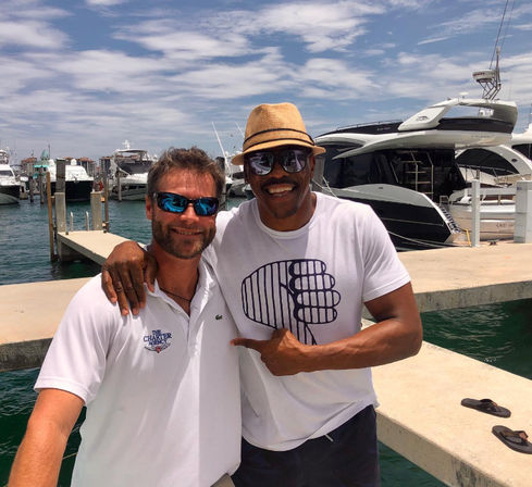 Two friends smiling on a sunny marina dock with luxury yachts in the background, one wearing a straw hat and both wearing sunglasses.