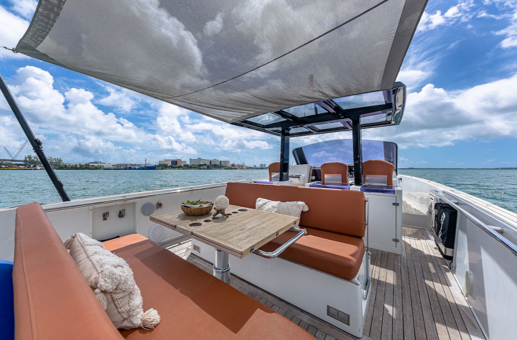 Sunlit yacht deck with orange bench cushions and wooden table under a shade sail, coastal skyline and blue sky over calm harbor water.