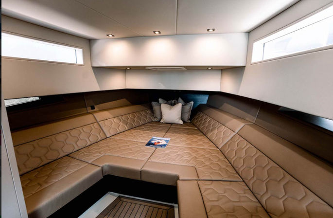 Cozy luxury yacht interior — v-berth boat cabin with tan quilted leather seating forming a triangular bed, side windows, recessed ceiling lights and decorative pillows.