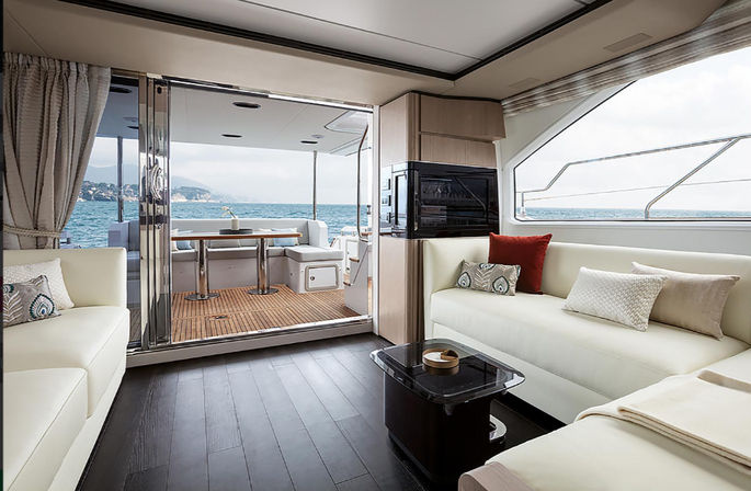 Bright modern yacht salon with cream leather sofas, colorful throw pillows and glass coffee table, sliding doors opening to a teak aft deck dining area and open ocean with distant coastline.