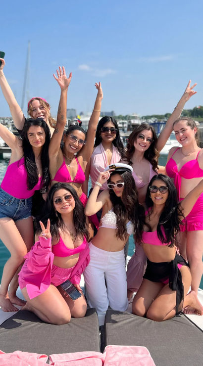 Group of friends in bright pink swimsuits cheering on the bow of a yacht at a sunny marina with docked boats and clear blue sky, summer boat party vibe
