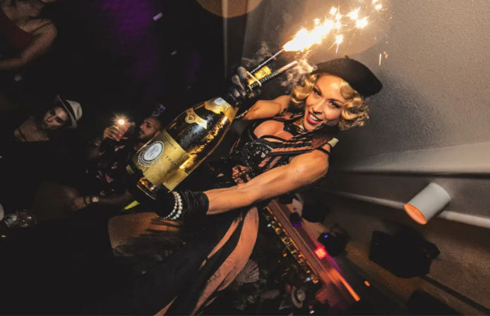 Glamorous performer in a beret holding a giant champagne bottle topped with sparklers, celebrating above a lively nightclub crowd with flashing phones.