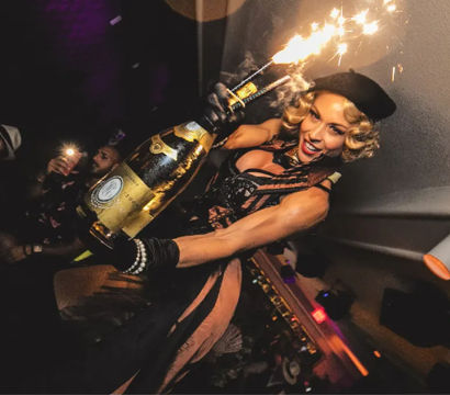 Glamorous performer in a beret holding a giant champagne bottle topped with sparklers, celebrating above a lively nightclub crowd with flashing phones.