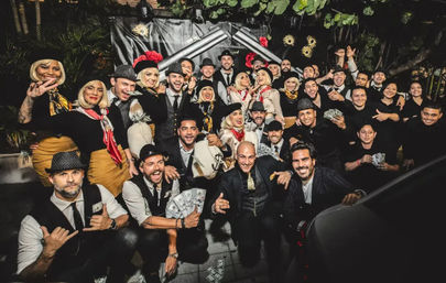 Large costumed group at an outdoor nighttime 1920s-style party, smiling and posing with fedoras, vintage outfits and stacks of prop money