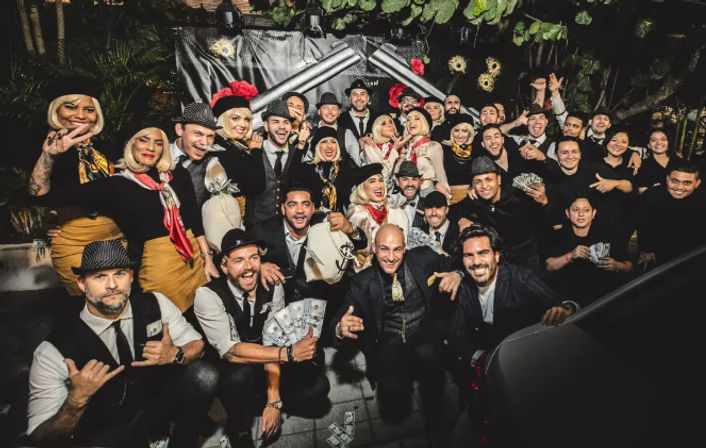 Large costumed group at an outdoor nighttime 1920s-style party, smiling and posing with fedoras, vintage outfits and stacks of prop money