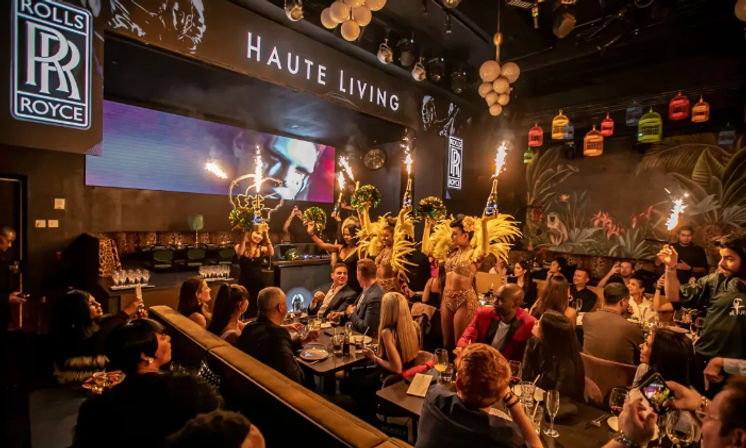 Upscale nightlife lounge with VIP tables, showgirls in yellow feather costumes carrying sparking bottle service as diners celebrate around candlelit tables under colorful hanging lanterns