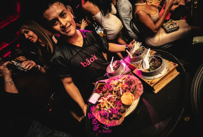 Smiling server in a neon-lit nightclub carrying a platter of colorful Asian-fusion small plates and skewers to a lively crowd