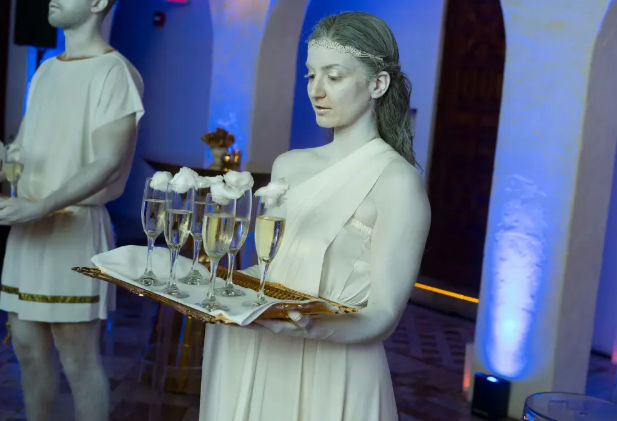 Living statue–style server in white toga holding a tray of champagne flutes topped with cotton 'foam' at a blue-lit indoor reception