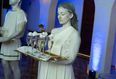Living statue–style server in white toga holding a tray of champagne flutes topped with cotton 'foam' at a blue-lit indoor reception