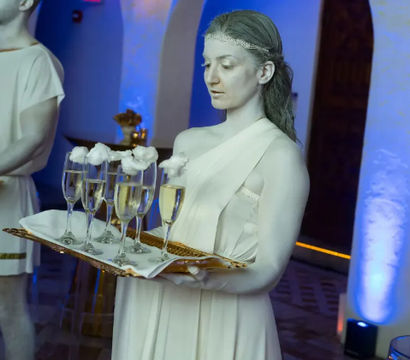 Living statue–style server in white toga holding a tray of champagne flutes topped with cotton 'foam' at a blue-lit indoor reception