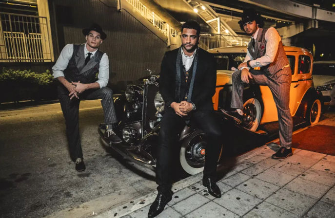Three sharply dressed men in retro suits and fedoras pose on and around a yellow vintage car with whitewall tires on a dimly lit urban street at night