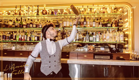 Flair bartender in vest and bow tie tossing a cocktail shaker above a marble bar counter in an upscale cocktail bar with mirrored shelves of liquor bottles and warm golden lighting.