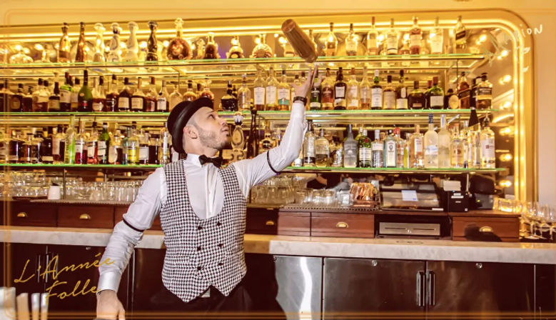 Flair bartender in vest and bow tie tossing a cocktail shaker above a marble bar counter in an upscale cocktail bar with mirrored shelves of liquor bottles and warm golden lighting.