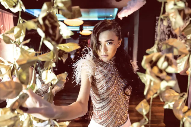 Glamorous portrait of a woman in a beaded, feather-shoulder dress reaching through hanging gold leaves in a dimly lit party venue.