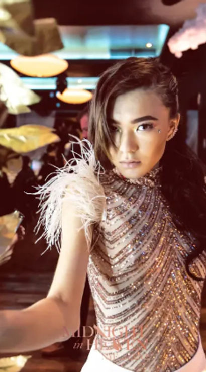 Glamorous portrait of a woman in a beaded, feather-shoulder dress reaching through hanging gold leaves in a dimly lit party venue.