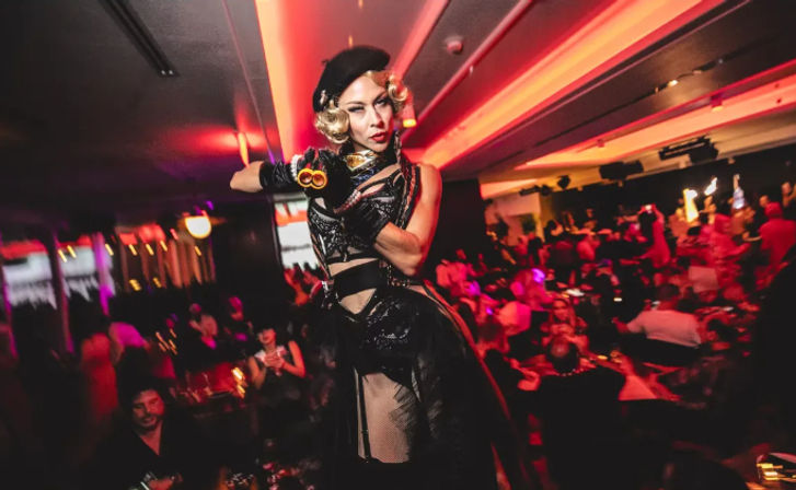Glamorous cabaret performer in black corset, beret and gloves striking a dramatic pose and pointing a prop toward the camera on a red-lit nightclub stage with a seated audience.