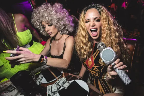 Two women at a lively nightclub VIP table — one with a curly silver wig checking her phone, the other with long wavy blonde hair holding a sparkly microphone and singing, dollar bills scattered on the table under purple party lights.
