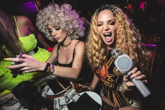 Two women at a lively nightclub VIP table — one with a curly silver wig checking her phone, the other with long wavy blonde hair holding a sparkly microphone and singing, dollar bills scattered on the table under purple party lights.