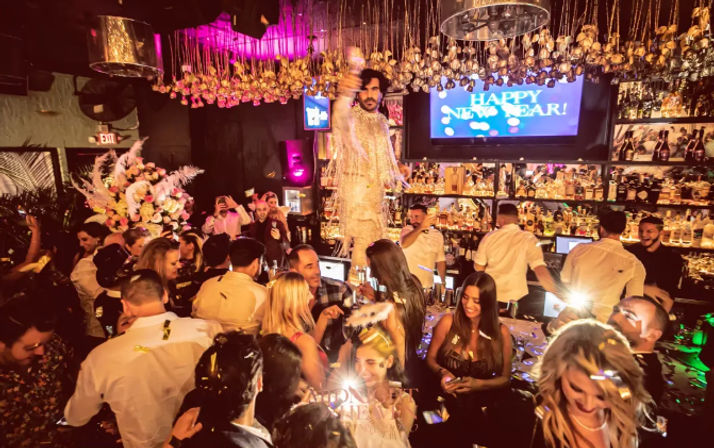 Packed nightclub New Year’s Eve party with guests celebrating as a glittering performer stands on the bar under hanging lights, confetti, floral arrangements and a 'Happy New Year' screen.