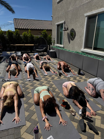 Outdoor yoga class on a sunny suburban tiled patio — group of people in child’s pose on gray mats with water bottles, sandals, a hanging wicker chair, palm tree and string lights in the background.