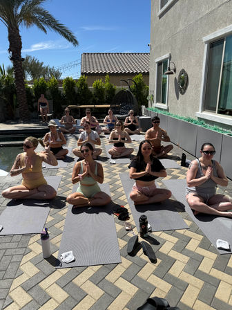 Sunny backyard poolside yoga class on a tiled patio with students seated on mats in meditation pose beneath a palm tree