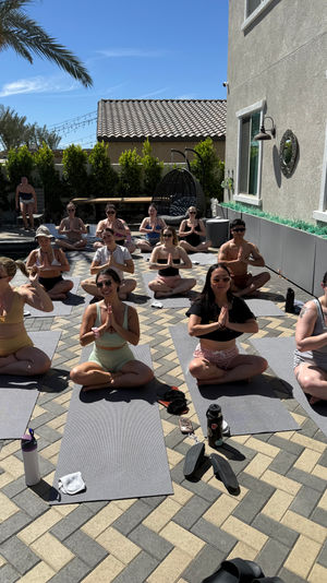 Sunny backyard poolside yoga class on a tiled patio with students seated on mats in meditation pose beneath a palm tree
