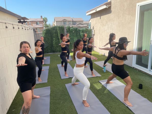 Group of women doing an outdoor yoga class on gray mats in a sunny backyard patio with artificial turf, all holding Warrior II poses