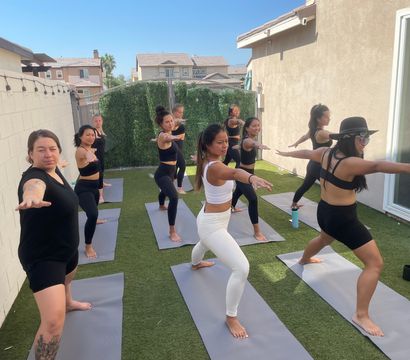 Group of women doing an outdoor yoga class on gray mats in a sunny backyard patio with artificial turf, all holding Warrior II poses