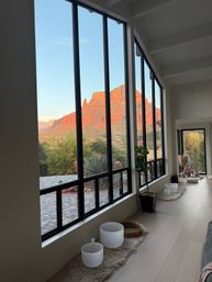 Sunset red-rock mesa and desert landscape framed by tall black-framed floor-to-ceiling windows of a modern Arizona home, interior with plants, rugs and meditation bowls